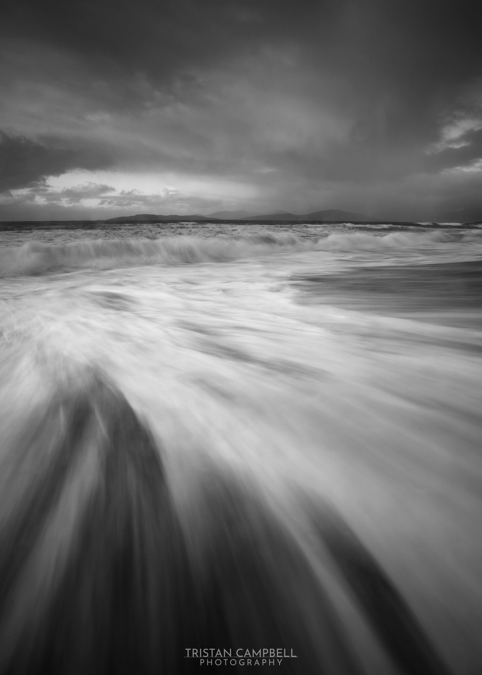 A monochrome seascape captures the drama of rushing waves under a turbulent sky. The water's motion blurs into silky streaks, contrasting with the crisp horizon where dark clouds hang over distant hills.