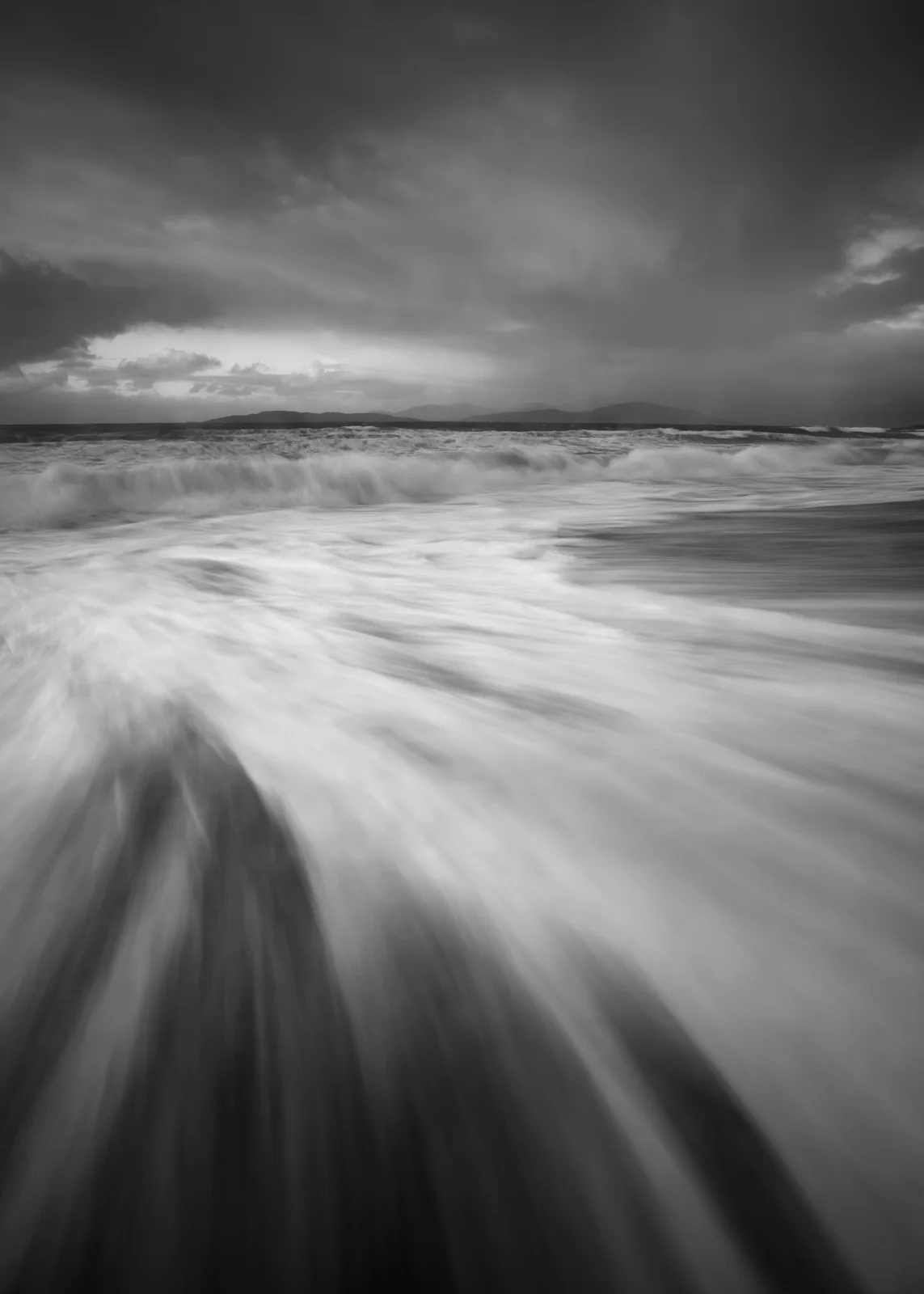 Moody monochrome seascape with foaming waves rolling onto sandy beach, distant mountains and stormy sky with break in clouds.