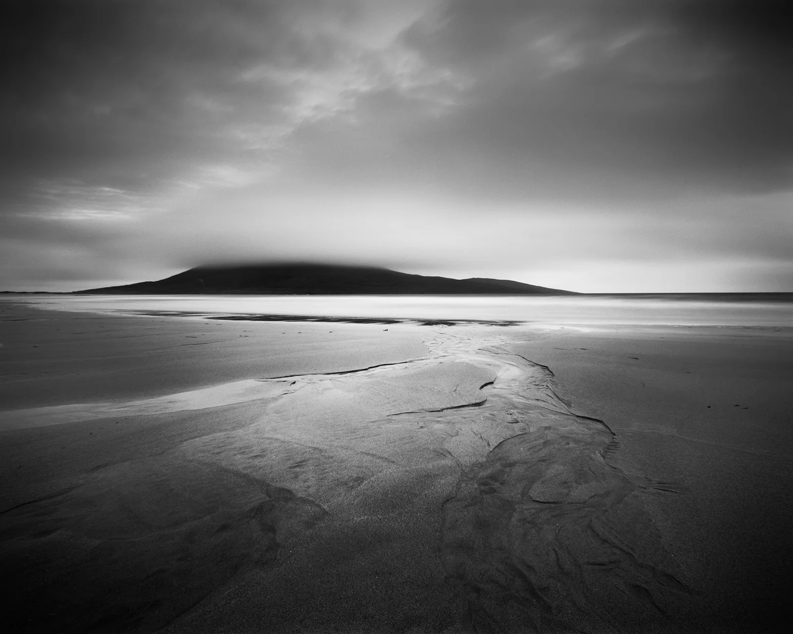 Black and white seascape with rippled sand flat in foreground, dark island silhouette across calm water, and overcast sky.