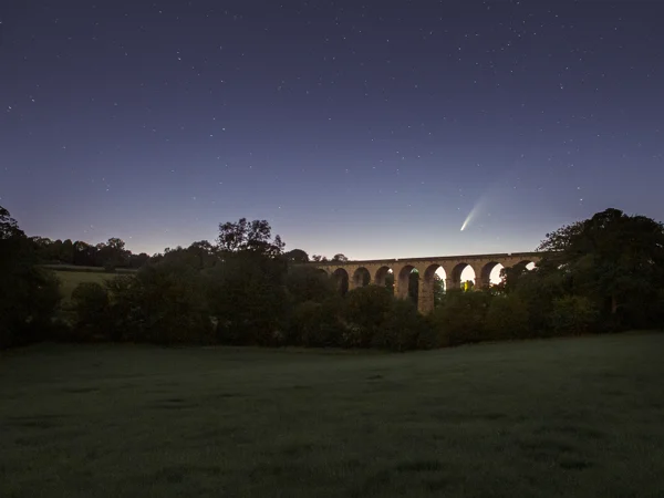 Comet Neowise above a stone railway viaduct in a wooded rural valley under a starry twilight sky