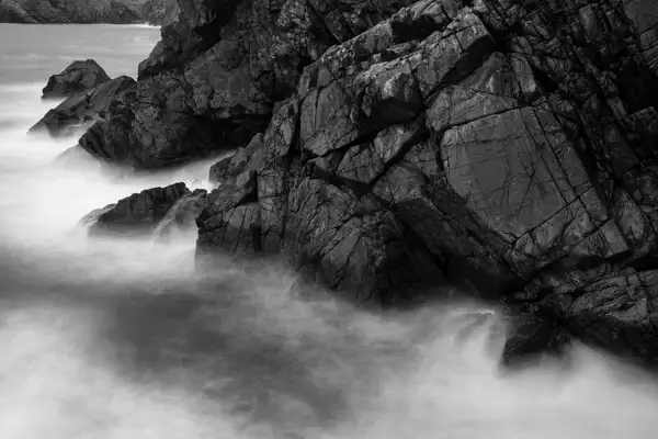 Rocky shoreline with rugged, dark stones sharply jutting into the sea. Waves crash and flow around the rocks, captured with a long exposure that creates a smooth, misty appearance in the water. The overall composition is in black and white, highlighting the textures and contrasts.