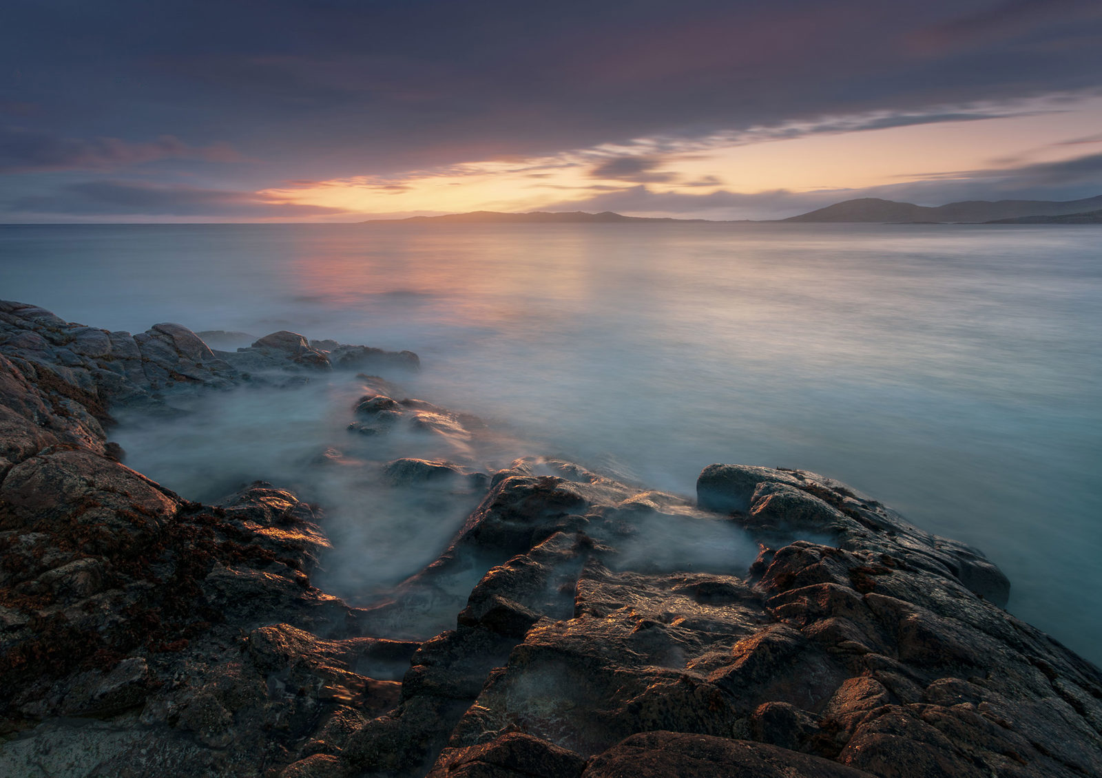 Rocky shoreline at sunset with smooth, misty waves lapping against the rocks. The sky is painted in hues of orange, pink, and purple, reflecting softly on the calm sea. Hills are faintly visible on the horizon under a dramatic sky.