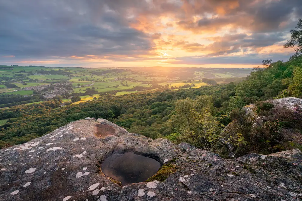 Sunset over a verdant landscape viewed from a rocky cliff. The sky is a blend of orange and purple hues as the sun sets above the horizon. A small pool of water is nestled in the foreground on the rock surface, reflecting the sunset. Below, dense forests stretch out towards patchwork fields and a distant village, bathed in warm golden light.