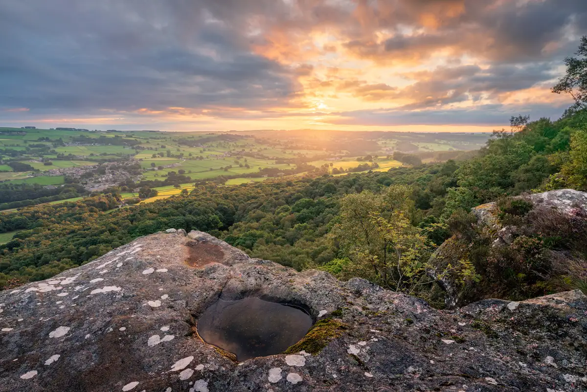 Sunset over a verdant landscape viewed from a rocky cliff. The sky is a blend of orange and purple hues as the sun sets above the horizon. A small pool of water is nestled in the foreground on the rock surface, reflecting the sunset. Below, dense forests stretch out towards patchwork fields and a distant village, bathed in warm golden light.