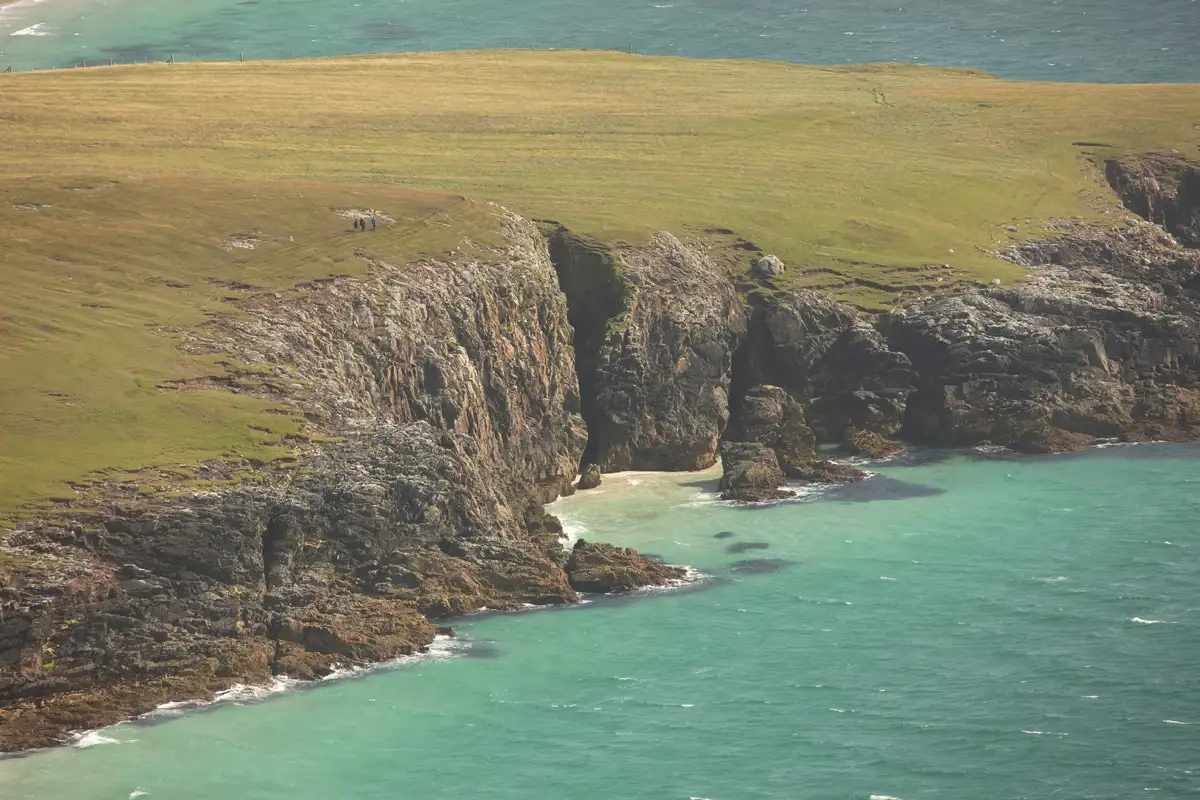 Rocky coastline with steep cliffs meeting turquoise sea. The grassy headland above the cliffs stretches out into the distance. A small, secluded sandy cove is nestled between the cliffs. The sea is slightly wavy, and a few people are visible walking along the clifftop.