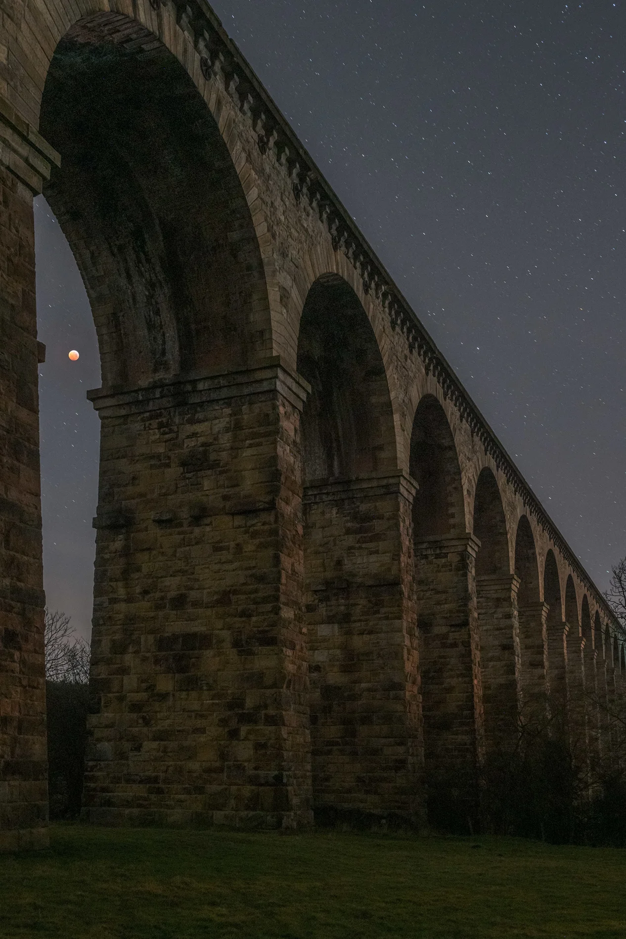 Stone railway viaduct arches at night under a starry sky with a glowing blood moon rising beside a pier