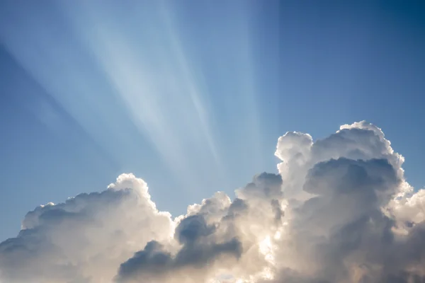 Crepuscular sun rays bursting behind towering white cumulus clouds against a deep blue sky