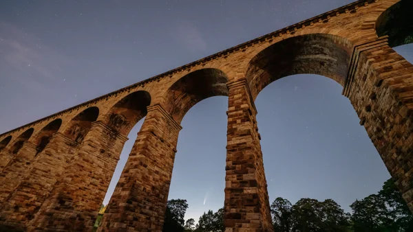 Floodlit stone railway viaduct arching across a starry night sky with Comet Neowise low above the trees