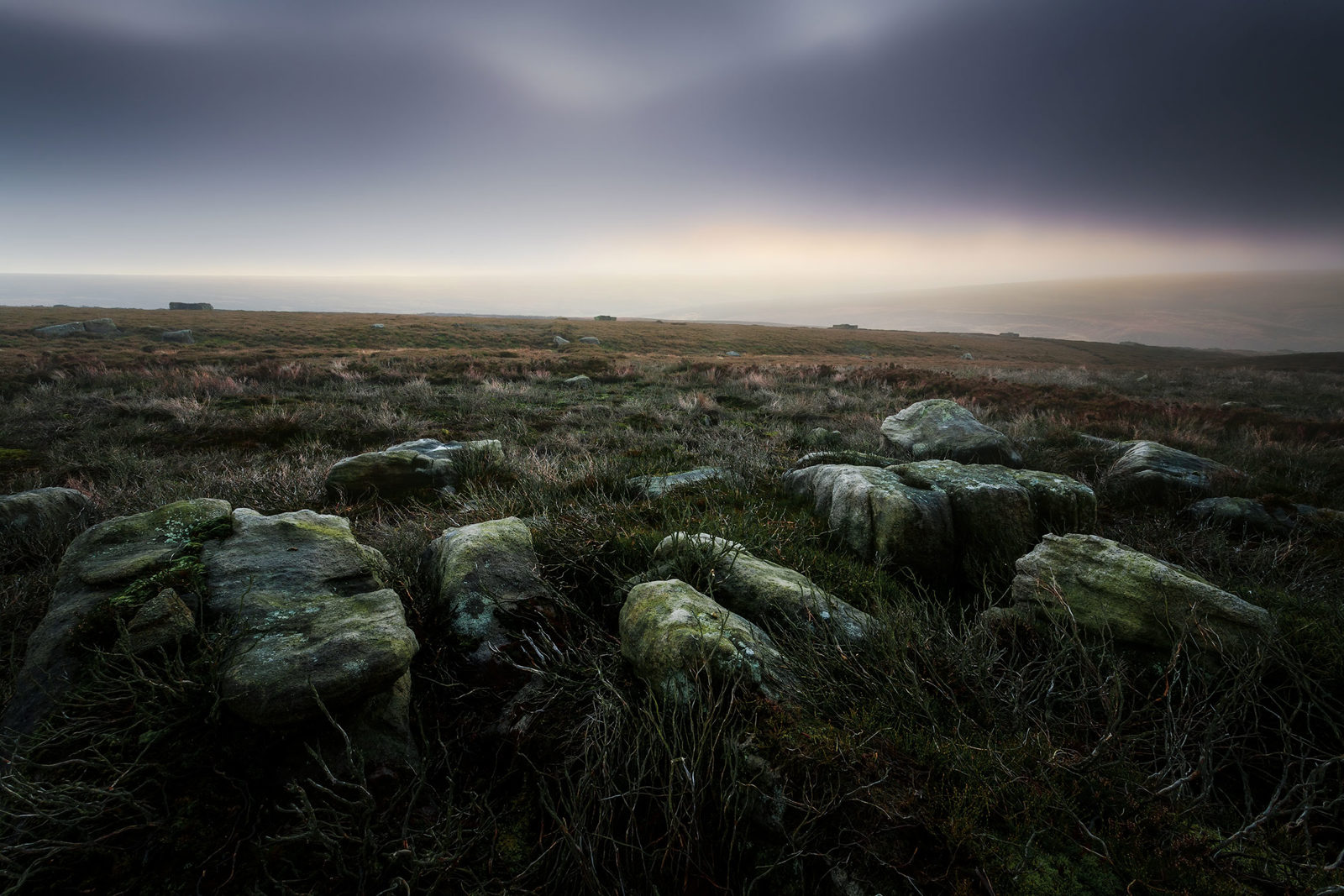 Misty moorland landscape with scattered large rocks in the foreground, covered in moss and surrounded by dried grasses. The horizon is shrouded in a soft, diffused light under a grey, overcast sky, creating a serene, atmospheric scene.