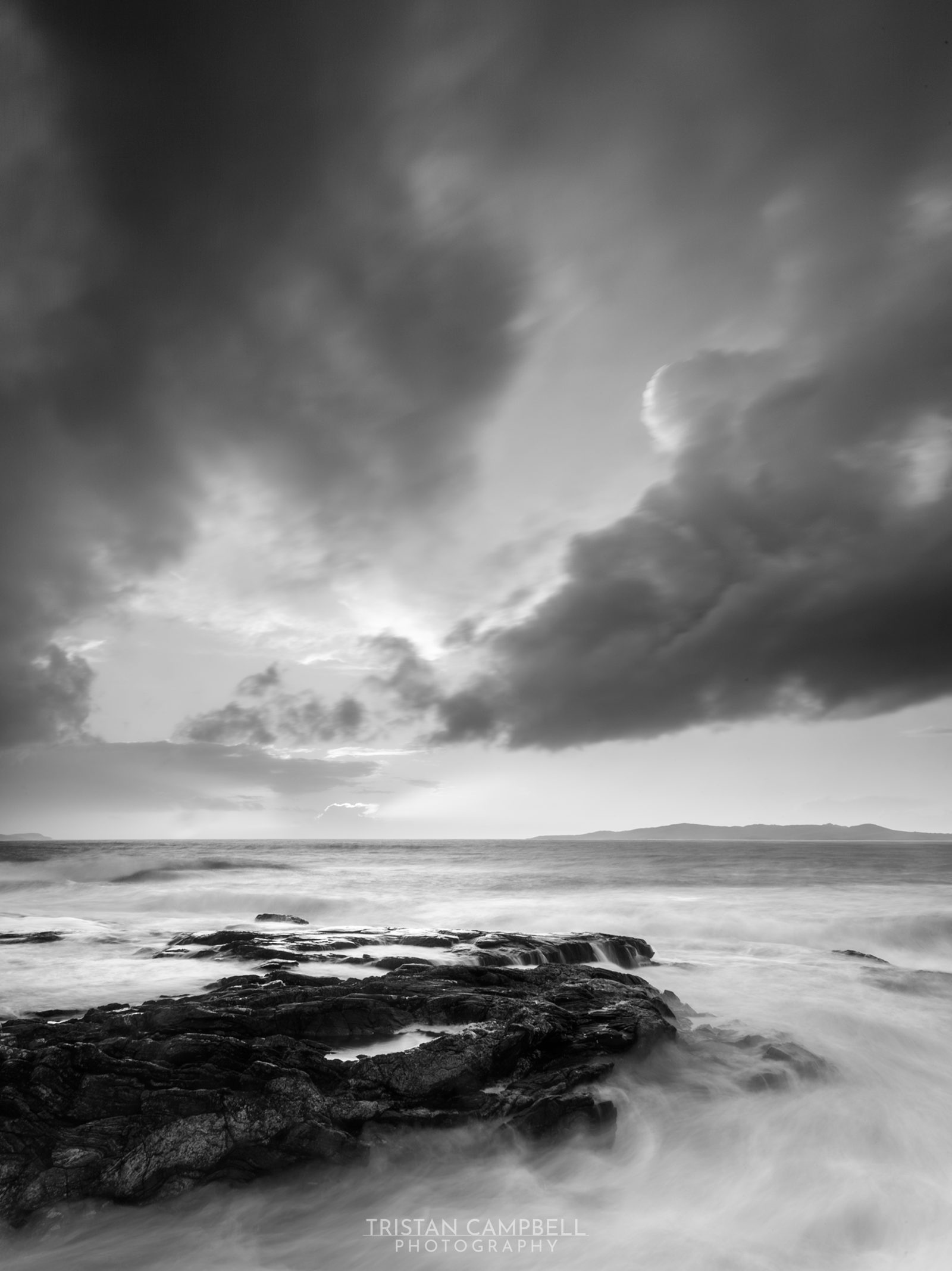 A monochrome seascape portrays tumultuous waves crashing against rugged rocks. Overhead, a dramatic sky with a blend of dark and light clouds suggests an emotive, changing weather pattern, with rays of sunlight piercing through in the distance.