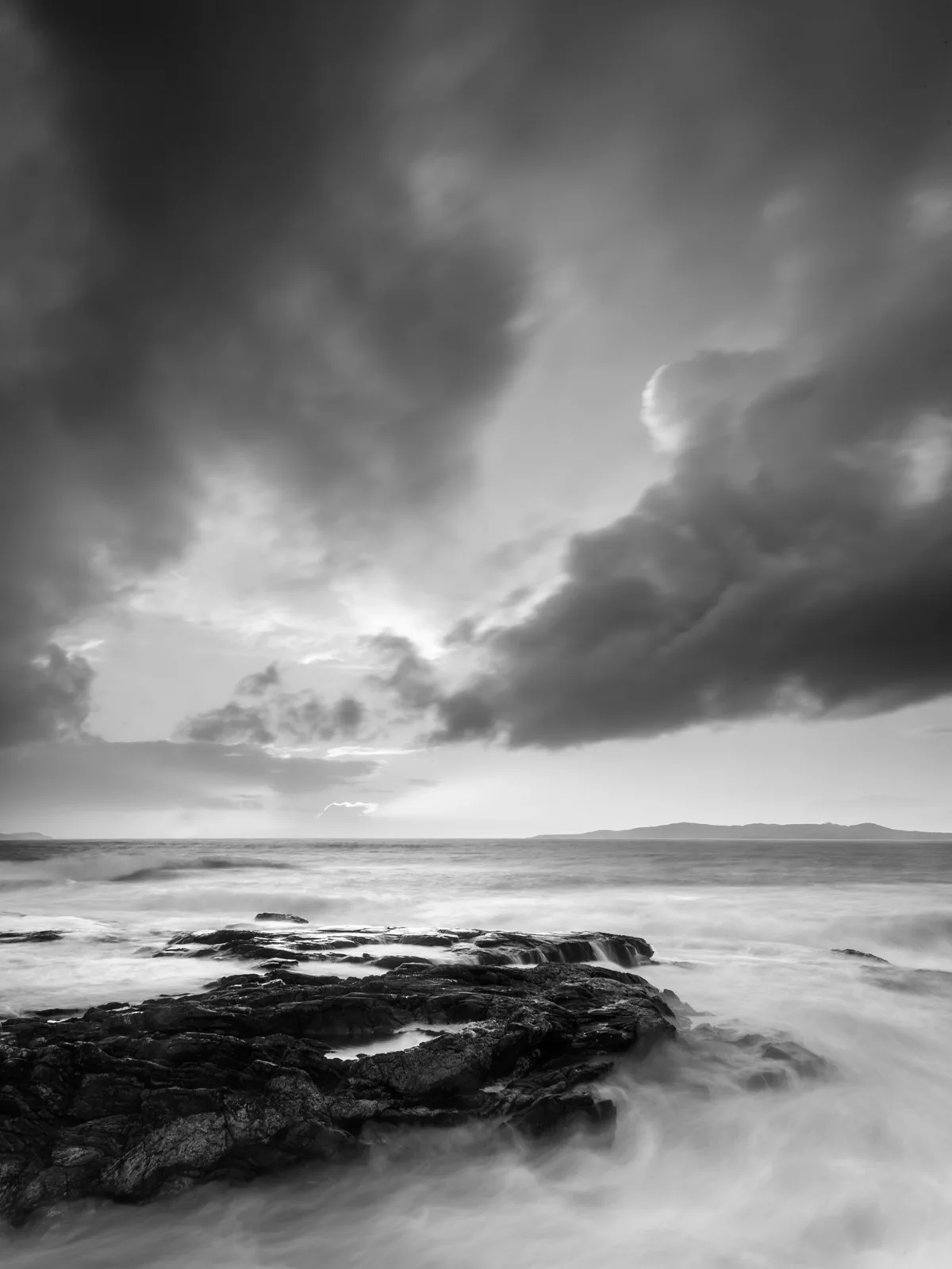 Black and white coastal landscape with dark rocky foreground, turbulent sea, distant hills, and dramatic cloudy sky with bright gap.