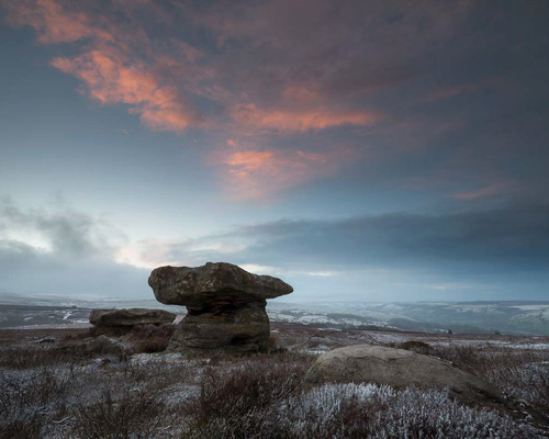 Moorland Landscapes: A distinctive rock formation resembling a mushroom under a dramatic sky on North Yorkshire moorland. Pink clouds hint at sunrise or sunset, gently contrasting with the muted blues of a vast, frost-kissed landscape. The terrain is rugged, dotted with low vegetation, suggesting cold, open wilderness. a person standing on a rocky beach