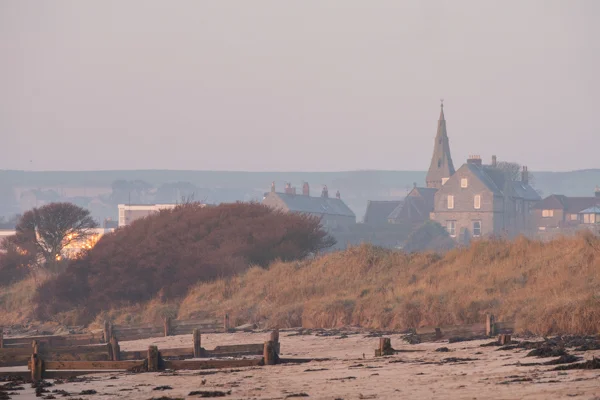 Church spire and stone houses behind misty coastal dunes and a sandy beach with weathered groynes at dawn.
