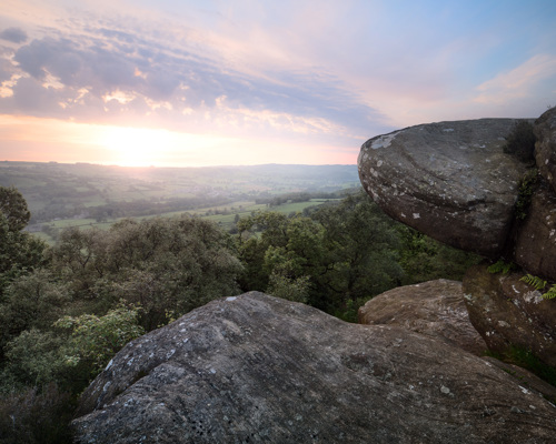 Moorland Landscapes: Large, weathered rock formations, covered with lichens, dominate the foreground while a lush canopy of trees spreads out below. The sky is painted with soft pastel hues as the sun dips near the horizon, casting a warm glow over the tranquil, rolling countryside. a view of a valley with trees and a sunset