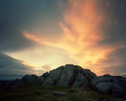 Moorland Landscapes: North Yorkshire's moors with an expansive and dramatic sky. Large, weathered rocks at the foreground create a rugged contrast against the sky's warm hues of orange and yellow merging with serene blues, suggesting a sunset. The long exposure has softened the clouds, enhancing the dreamlike atmosphere. a rocky landscape with a cloudy sky