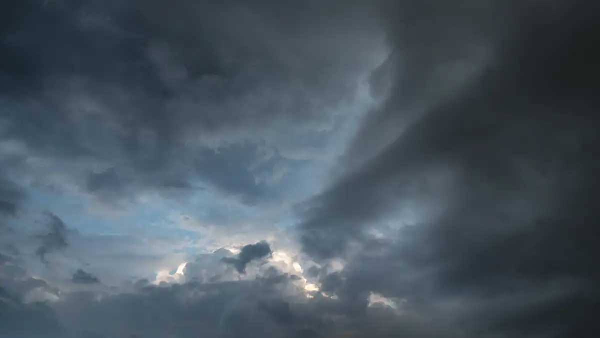 Dramatic cloudy sky with a mix of dark grey storm clouds and patches of lighter blue sky peeking through. Soft, diffused light highlights the edges of the clouds, creating a sense of depth and movement.