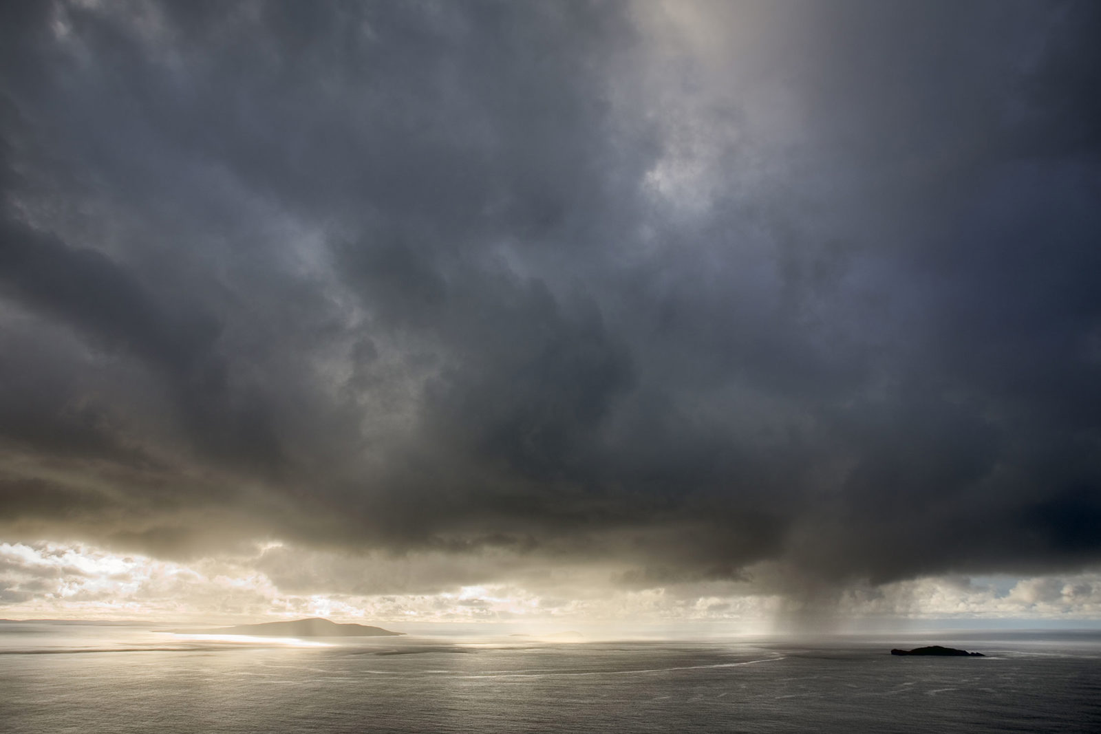 Dramatic cloudy sky over a vast ocean, with a dark storm cloud to the right pouring rain. Sunlight breaks through the clouds, illuminating a distant island and creating a stark contrast with the surrounding grey tones.