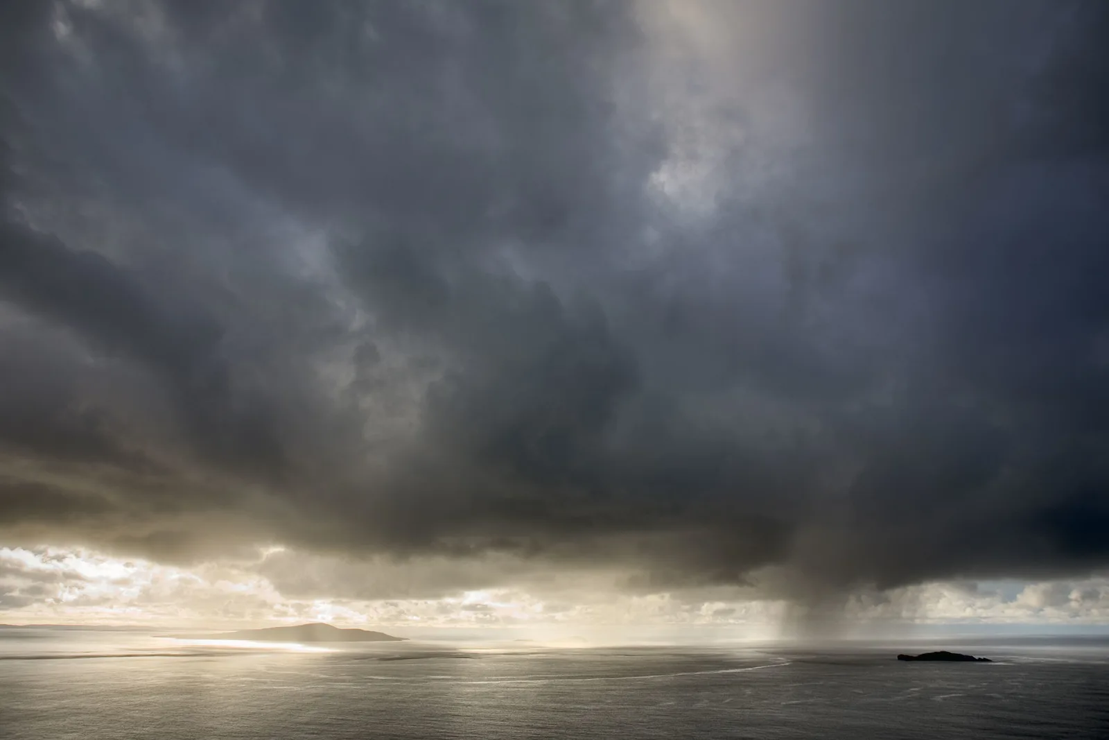 Dramatic coastal seascape with dark storm clouds towering over calm grey water and distant mountains under brooding sky.