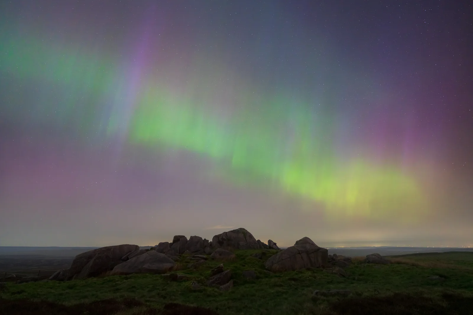 Northern lights dancing green and purple above dark rocky outcrops on moorland at night, scattered with stars.