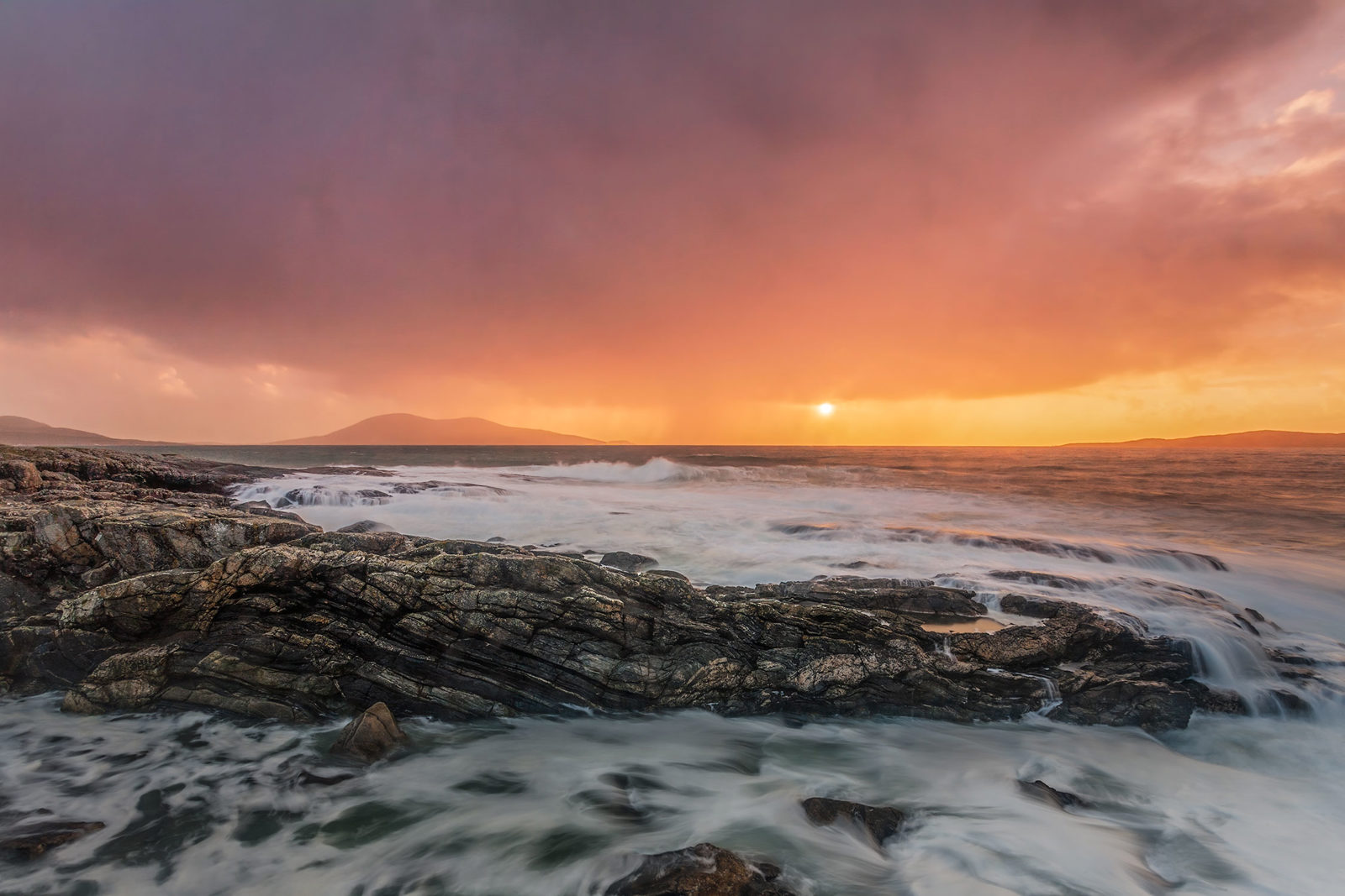 Rocky coastline with waves crashing against the shore under a dramatic sunset sky. The sky transitions from dark purple clouds to vibrant orange near the horizon, where the sun is partially visible. Distant hills are silhouetted by the setting sun.