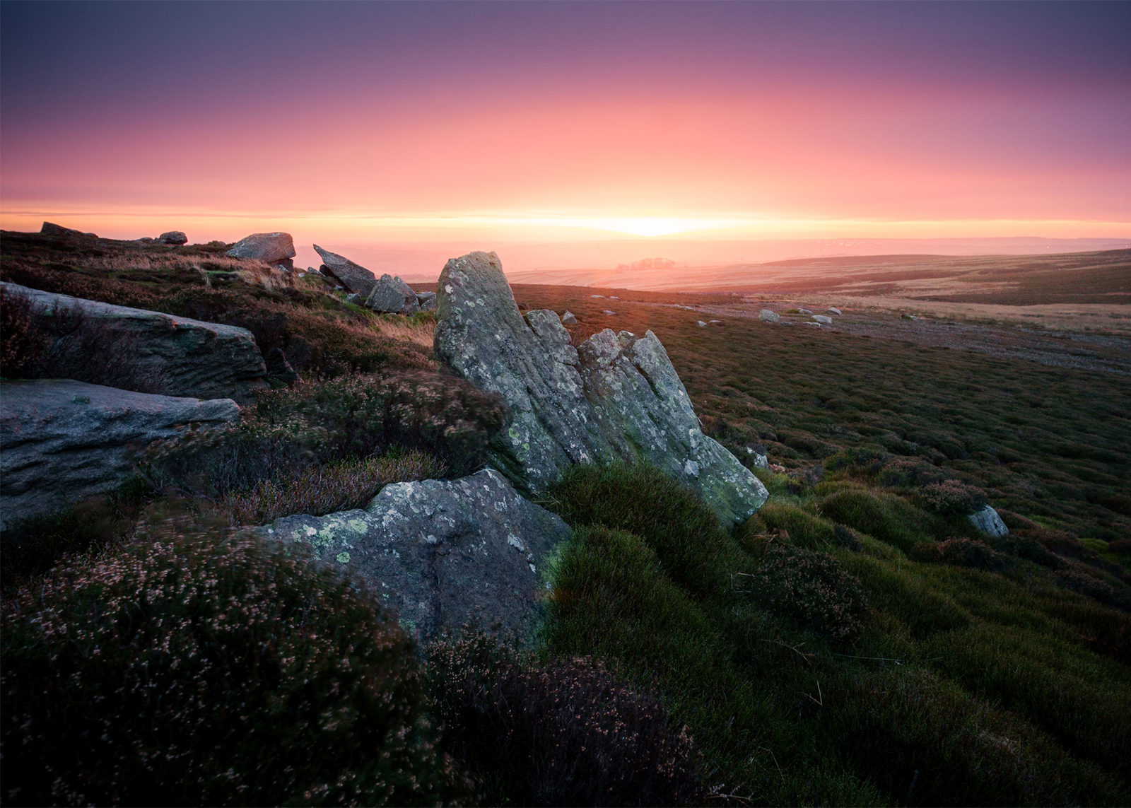 Sunrise over a moorland landscape, featuring large, weathered rocks in the foreground with patches of green and brown heather. The sky is a gradient of deep purple to warm orange, casting a soft glow across the rolling hills in the background.