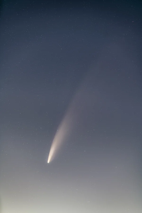 Comet NEOWISE with bright tail streaking across a star-filled twilight sky above a glowing horizon