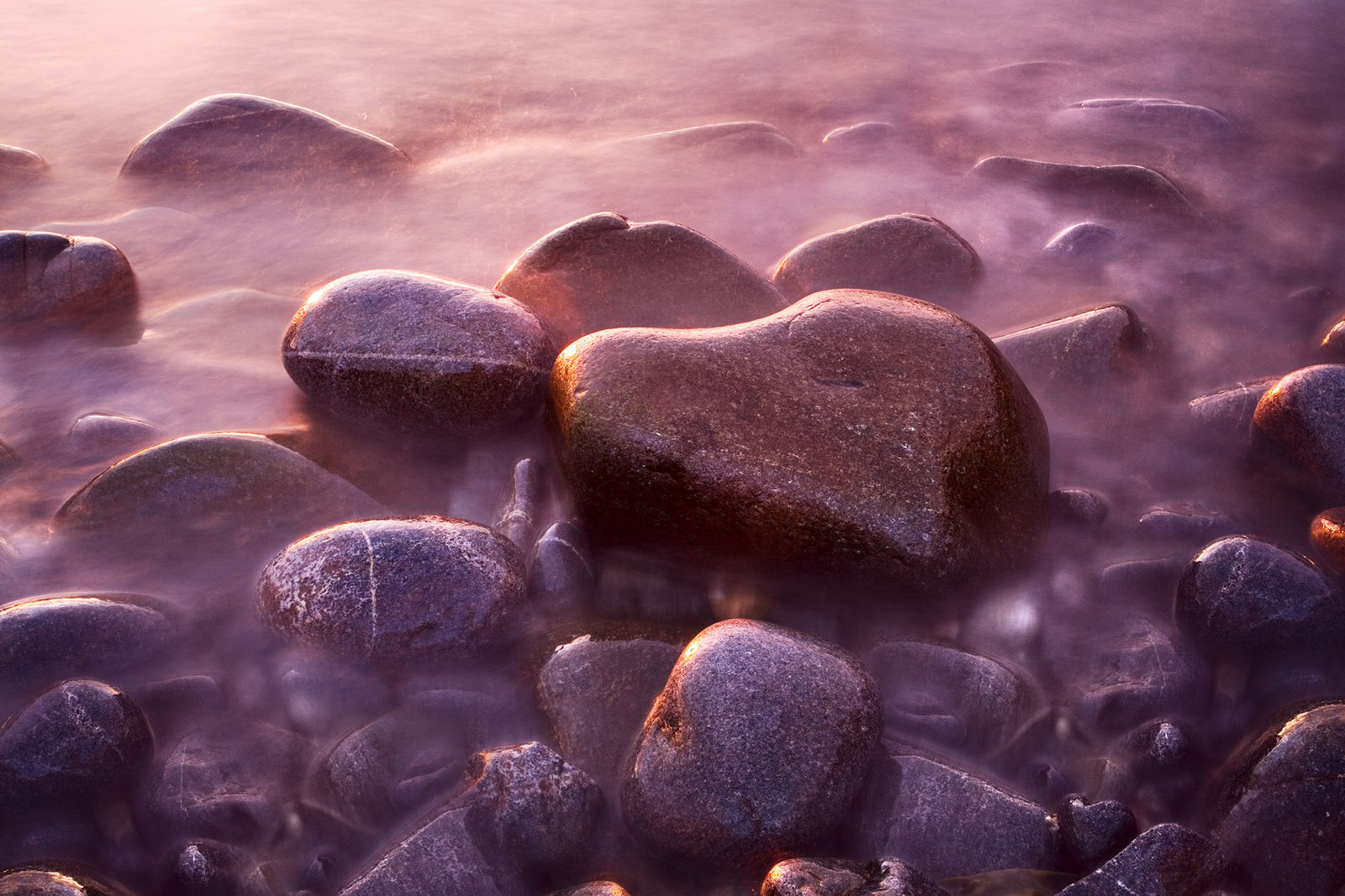 Smooth, rounded stones resting in shallow water, bathed in soft, warm light. A heart-shaped stone is prominent, with the surrounding rocks partially submerged in a gentle mist. The scene is imbued with a serene, ethereal quality.