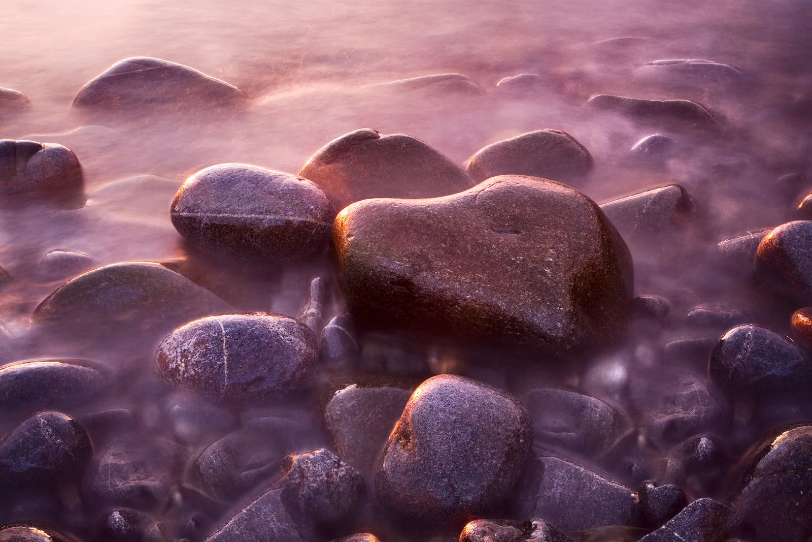 Smooth boulders emerging from misty pink water at dawn, with warm golden light illuminating their surfaces.