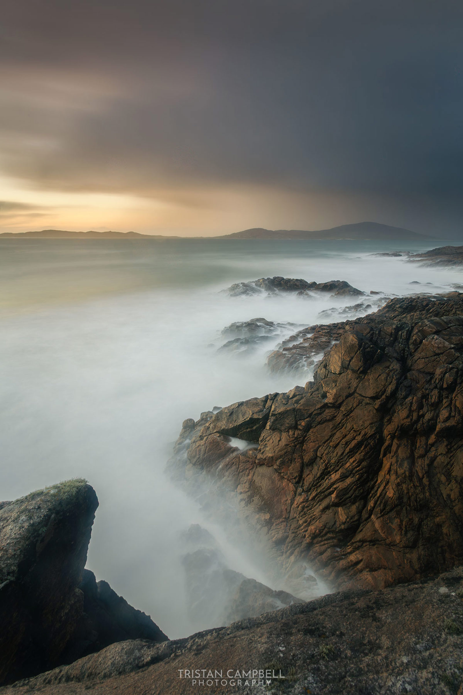 Rocky coastline at dusk or dawn with waves crashing against the rocks, creating a misty effect. The sky transitions from warm golden tones near the horizon to darker, stormy clouds above. Distant hills are faintly visible across a calm sea.