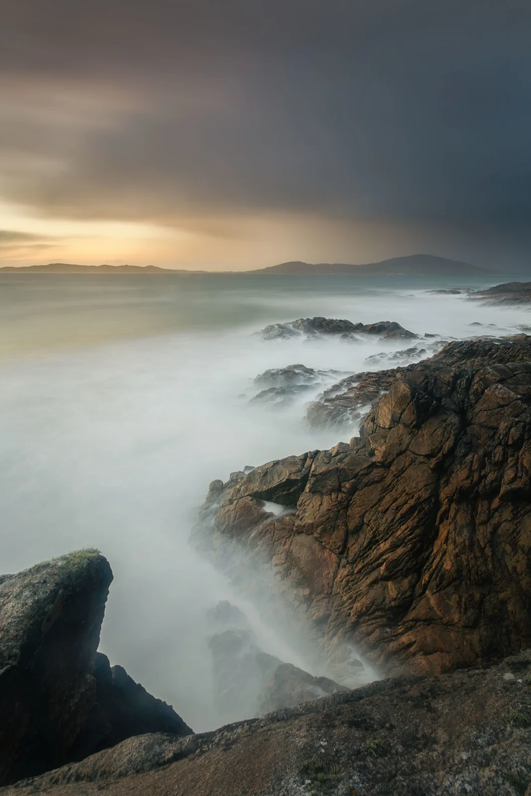Rocky granite coastline with long-exposure misty waves; distant mountains under overcast sky at dusk.