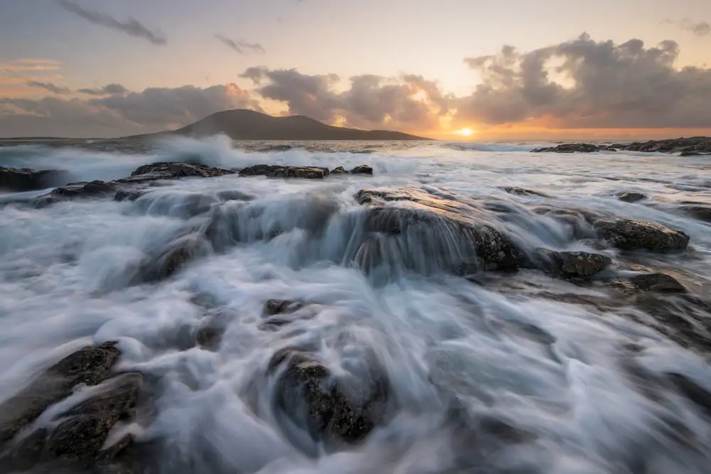 Waves cascade over dark, weathered rocks in the foreground, creating a misty, blurred effect. In the background, a softly illuminated horizon showcases a glowing orange sunset beneath a canopy of clouds. A distant silhouette of a hill rises on the left, partially obscured by the ocean spray.