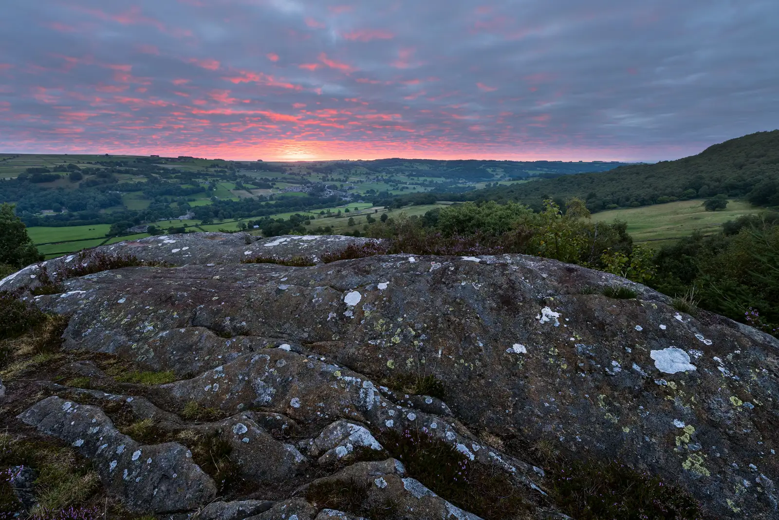 Rocky foreground with patches of lichen and small plants, overlooking a vast, lush green landscape of fields and rolling hills. A vibrant sunset casts pink and orange hues across a cloud-dappled sky, creating a dramatic horizon.