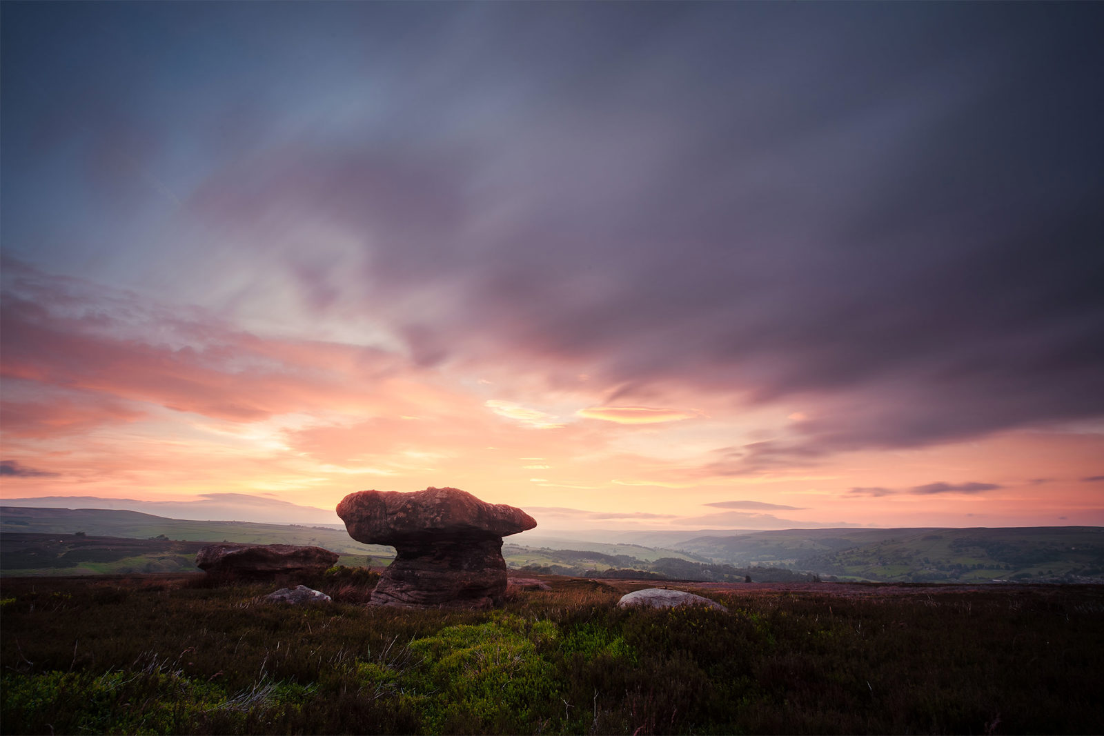 A prominent mushroom-shaped rock formation stands against a backdrop of rolling hills. The sky is filled with dramatic, fast-moving clouds painted in soft hues of purple, pink, and orange, suggesting a vibrant sunrise or sunset. The foreground features green and brown shrub-covered terrain.