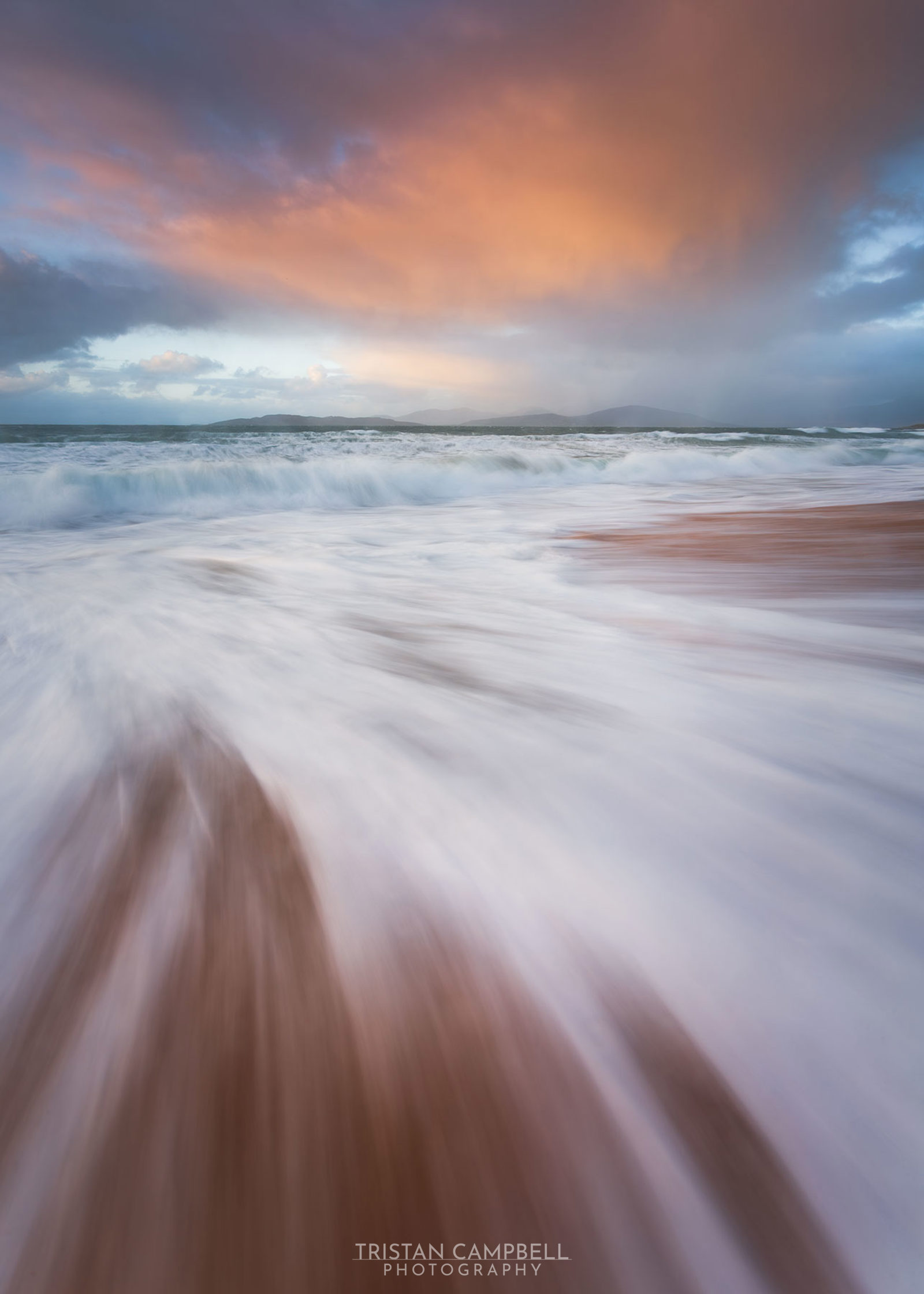 Waves gently sweep over a sandy beach under a dramatic sky. The sky is filled with vibrant orange and purple hues, casting a warm glow on the scene. The distant horizon displays rolling hills beneath a sky transitioning from blue to soft pink.