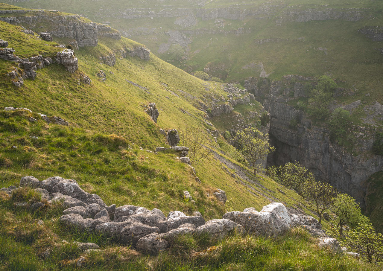 Sunlit rolling hills with patches of lush green grass and rocky outcrops. A steep rocky cliff drops into a shadowy gorge below, while scattered trees with fresh green leaves grow along the slope. The scene is bathed in soft, warm light, creating a serene and tranquil atmosphere.