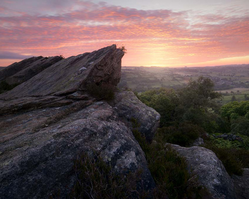 Moorland Landscapes: North Yorkshire vista at sunrise. Jagged rocks in the foreground overlook a patchwork of fields. The sky, a tapestry of pinks and oranges, fades to a gentle blue, signalling the start of a new day. a canyon with a mountain in the background