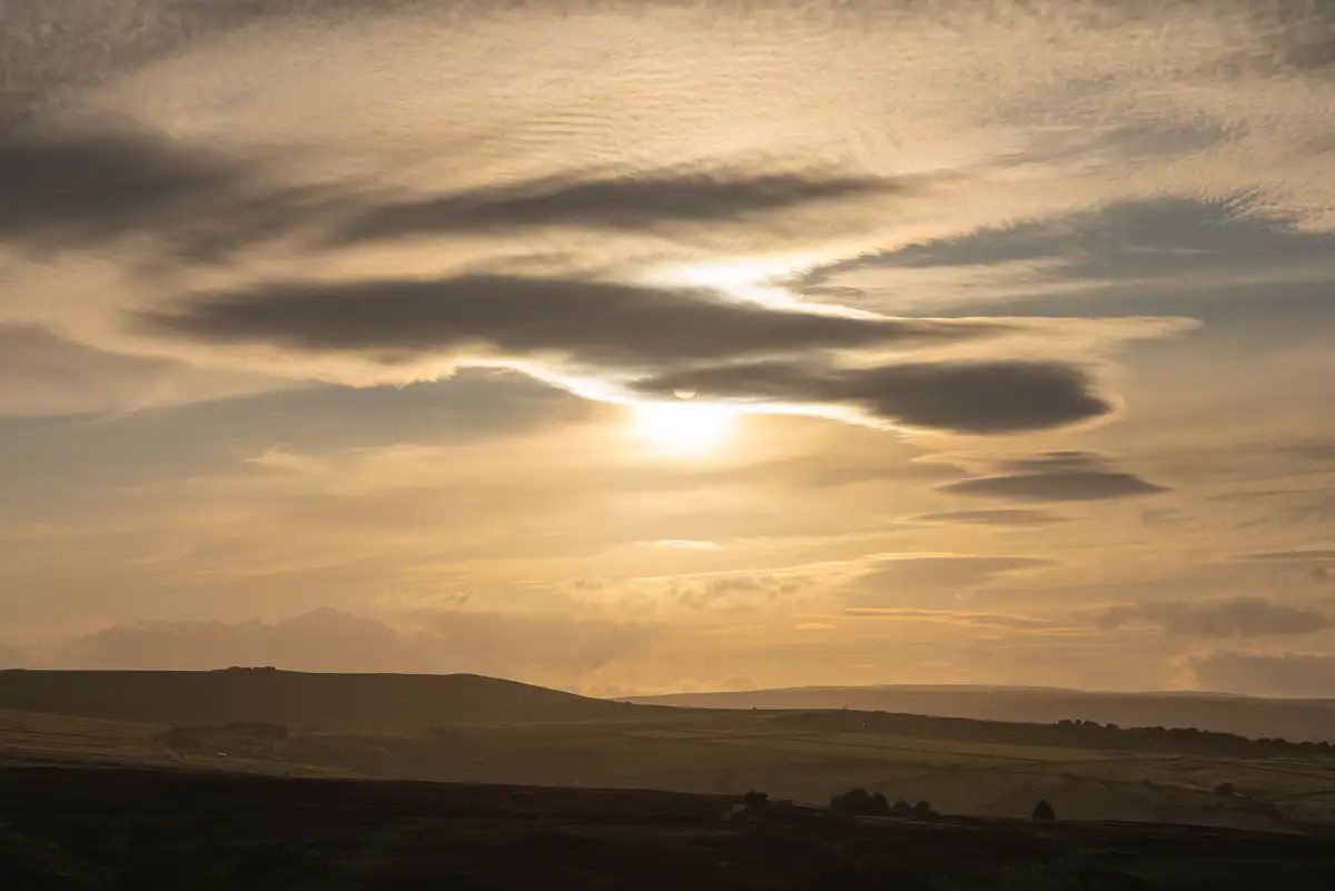 Golden sunrise with the sun partially hidden behind layered, wispy clouds. The landscape below features rolling, shadowy hills with scattered trees, creating a peaceful and expansive view.