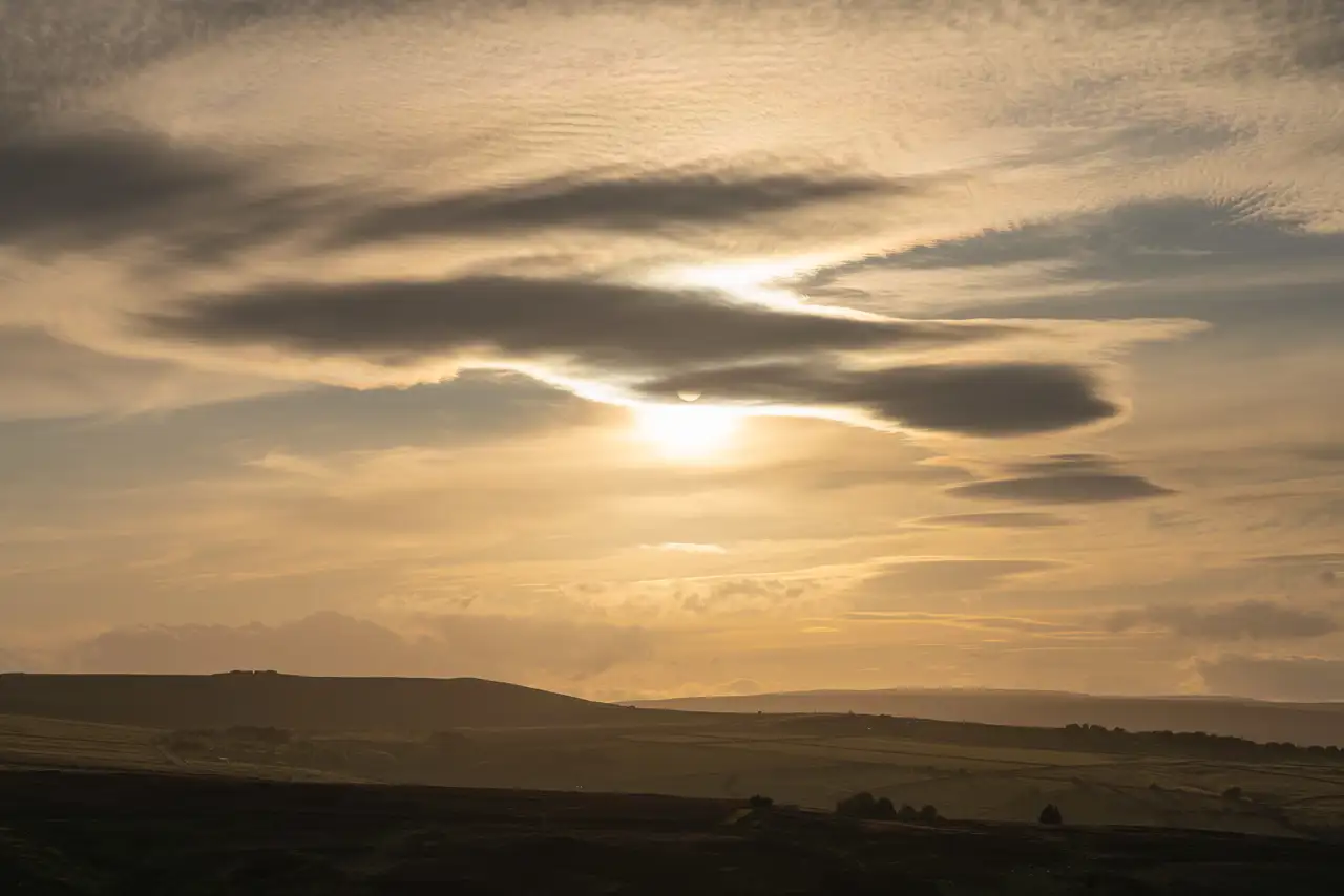 Golden sunrise with the sun partially hidden behind layered, wispy clouds. The landscape below features rolling, shadowy hills with scattered trees, creating a peaceful and expansive view.