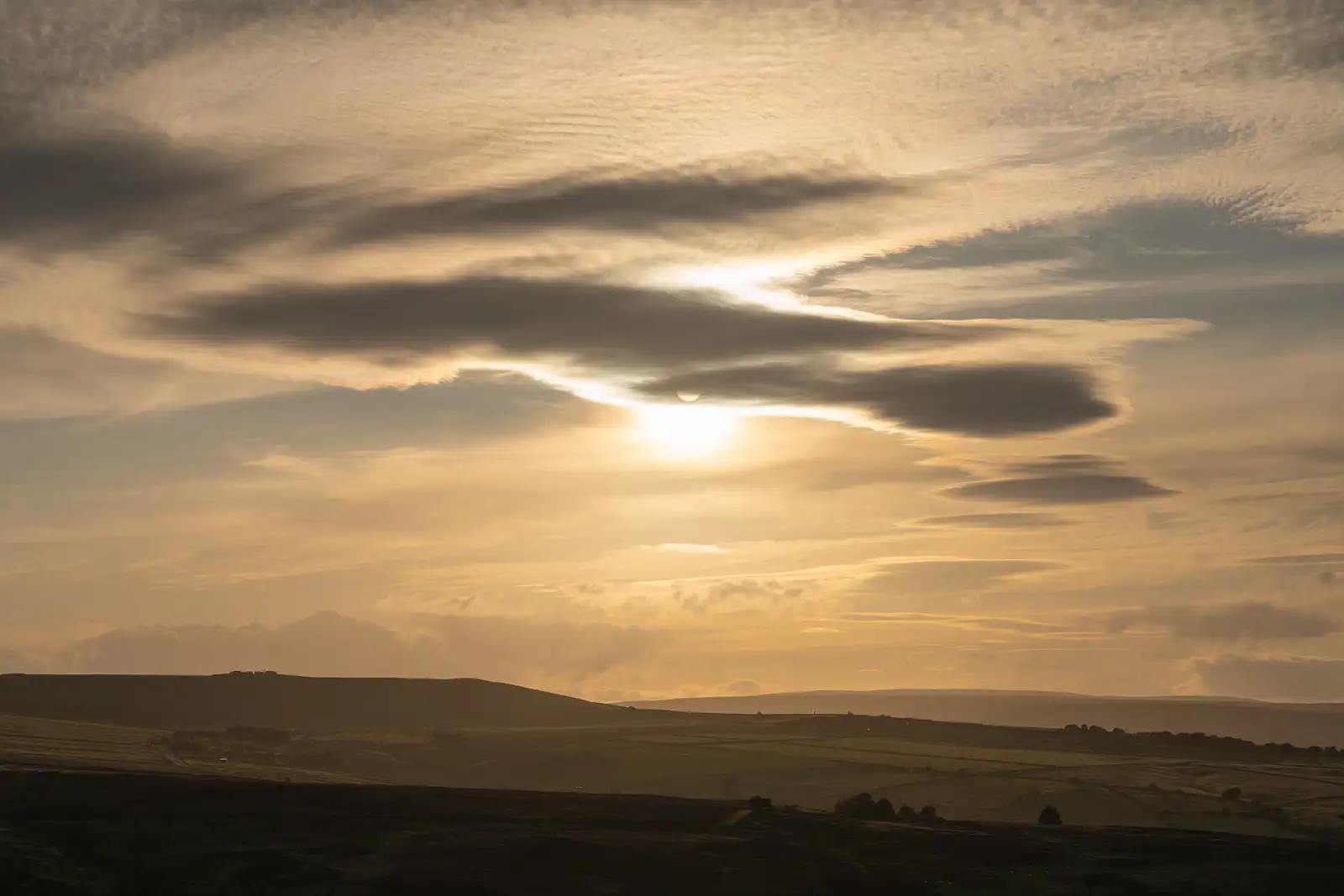 Golden sunrise with the sun partially hidden behind layered, wispy clouds. The landscape below features rolling, shadowy hills with scattered trees, creating a peaceful and expansive view.