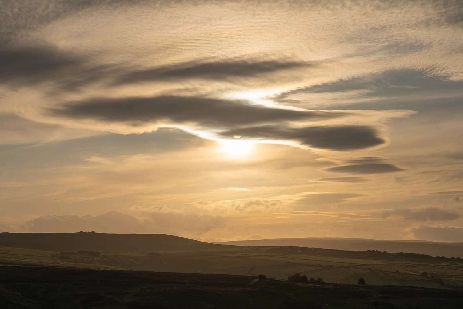 Sun breaking through lenticular clouds over rolling moorland hills at golden-hour sunset