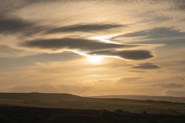 Sun breaking through lenticular clouds over rolling moorland hills at golden-hour sunset