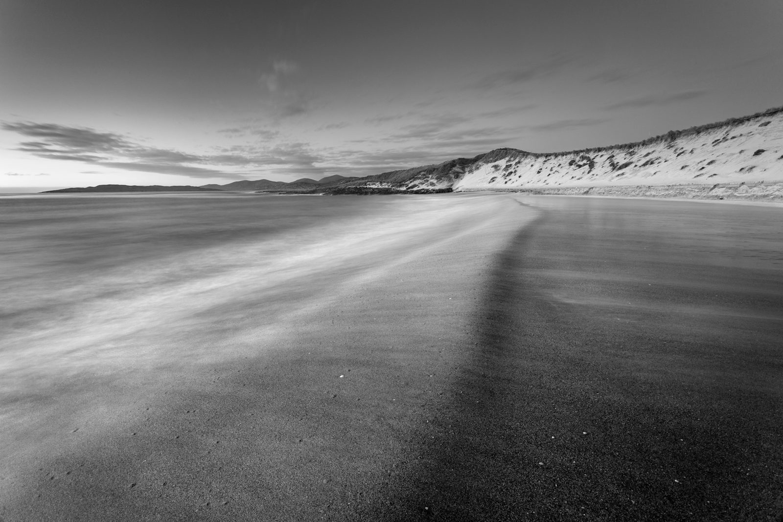 A monochrome landscape showcasing a vast beach with smooth sand leading into a soft blur of waves, under a sky with gentle cloud streaks. To the right, a series of dunes rise, peppered with sparse vegetation. In the distance, rolling hills meet the coastline, creating a serene vista.