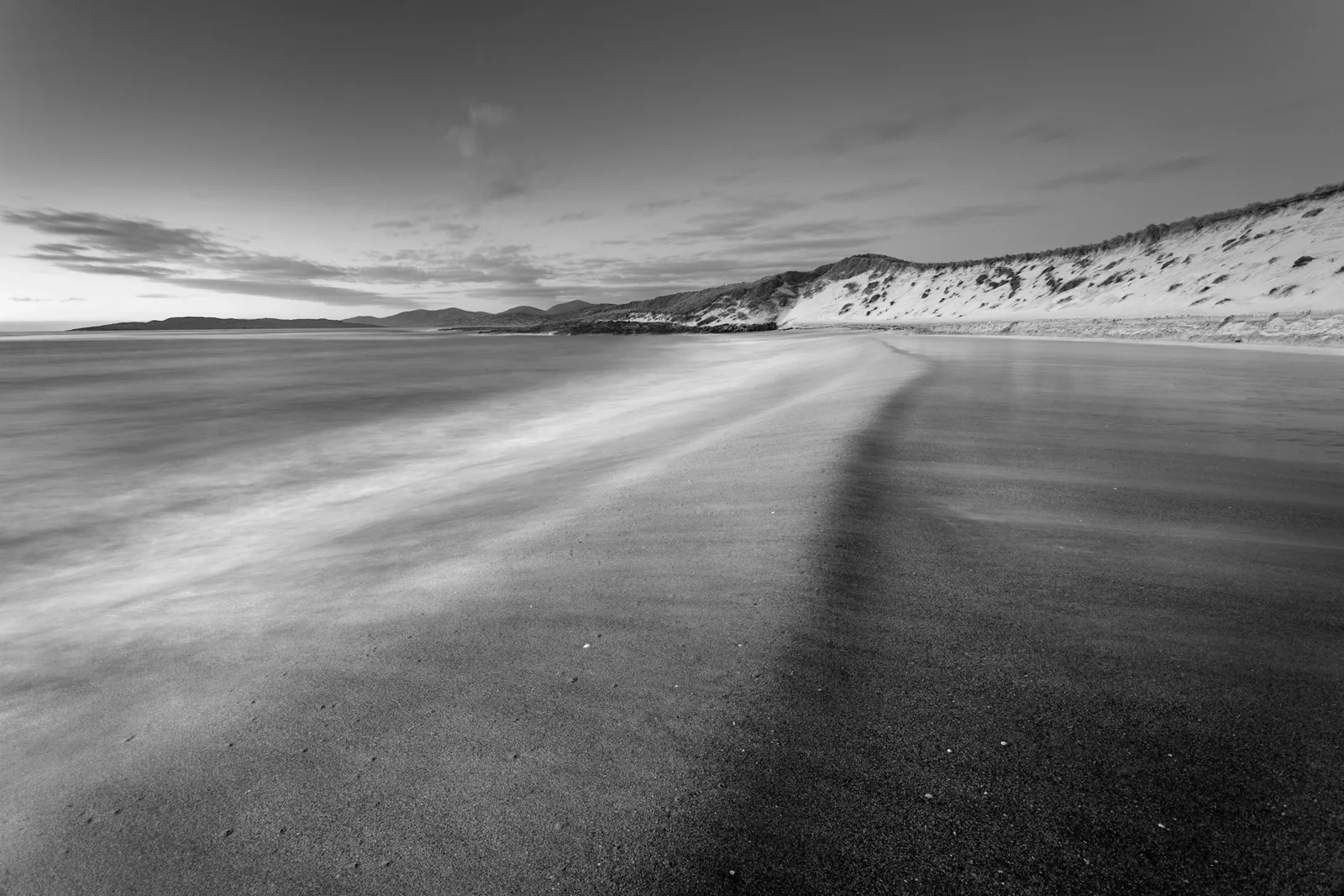 Black and white beach landscape with smooth sand, gentle waves, and snow-covered dunes beneath a grey sky.