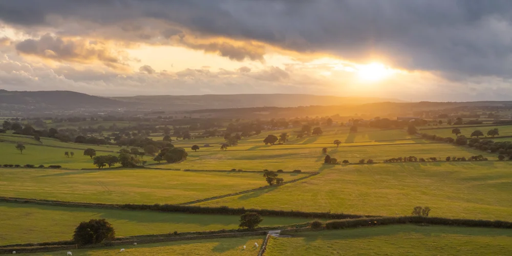 Patchwork of green fields and hedgerows in rolling countryside at sunset, with distant hills under a cloudy sky