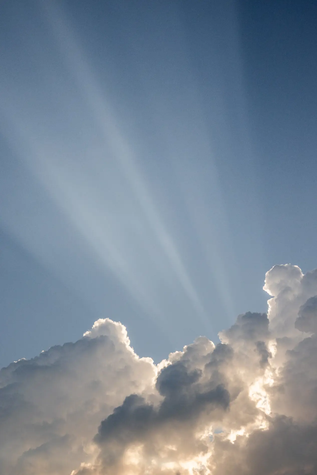 Crepuscular sun rays bursting through towering cumulus clouds against a deep blue summer sky