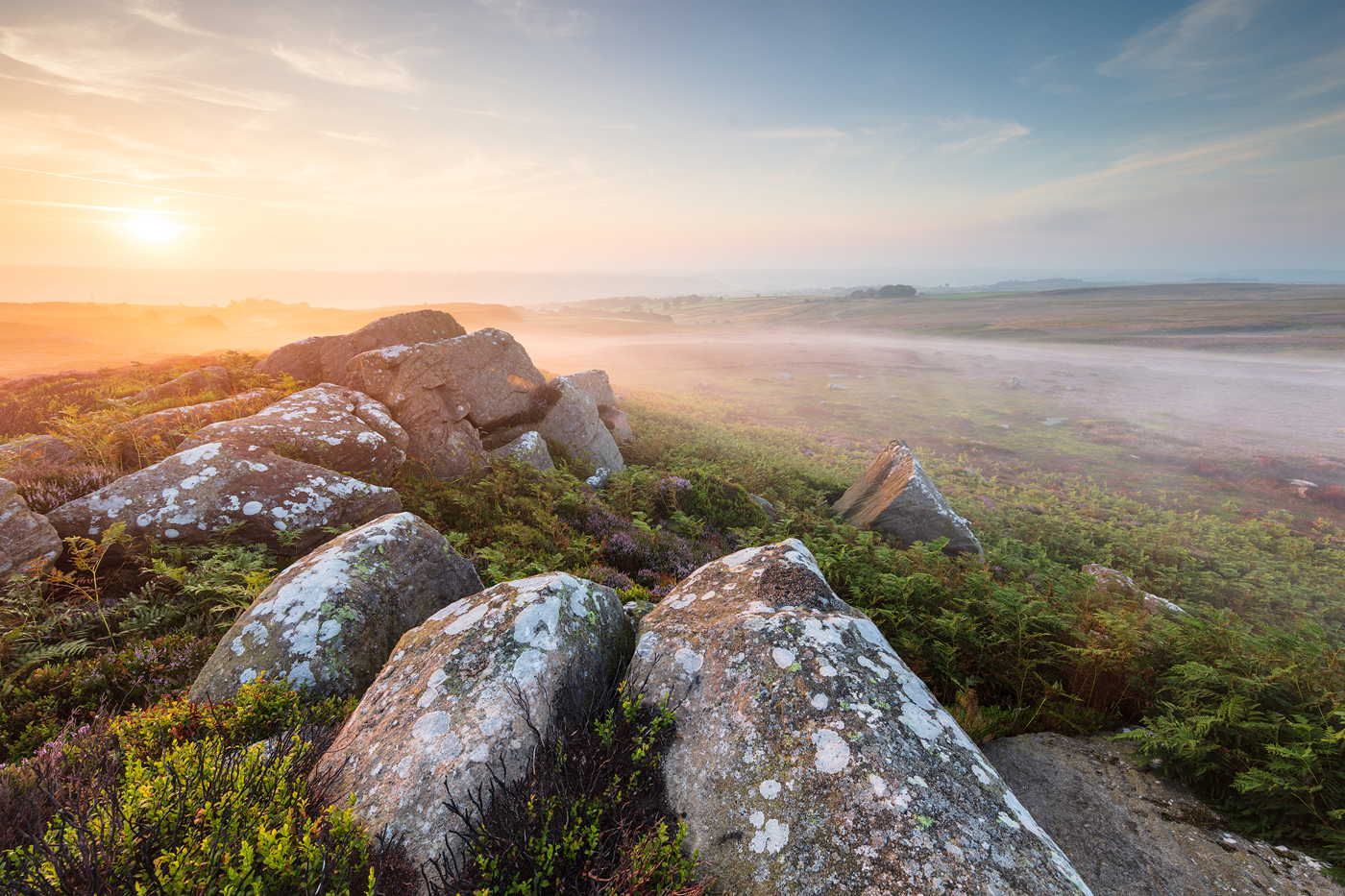 An intense sunrise bathes North Yorkshire moorland in soft light. Lichen-speckled rocks and heather streak the foreground, while a misty expanse stretches towards a hazy horizon. a rocky landscape with a valley in the background
