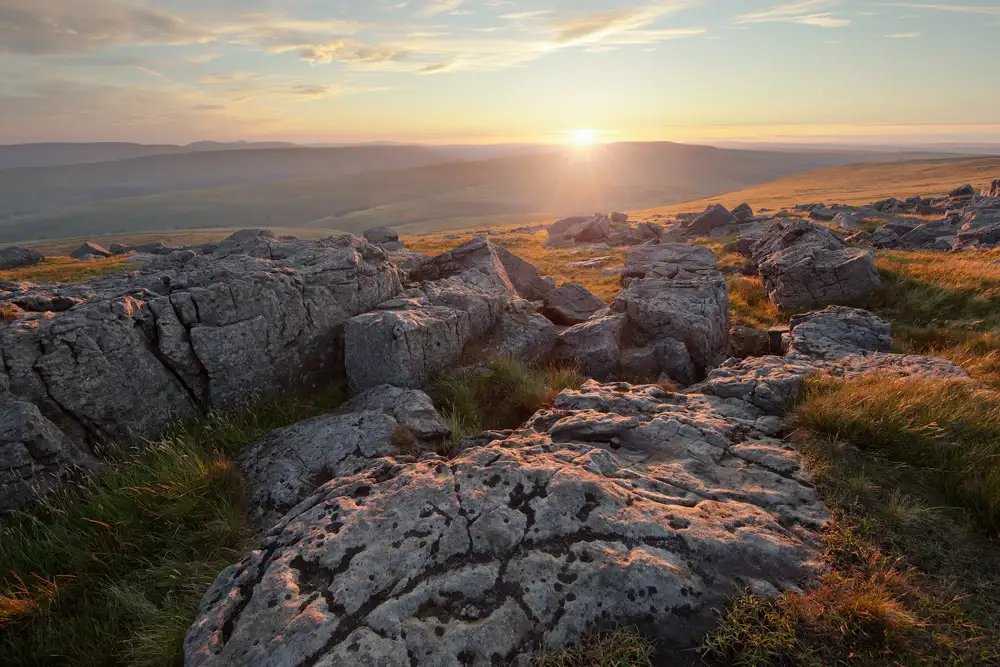 Rocky moorland terrain with large, weathered boulders illuminated by golden sunlight at sunset. Grassy patches with long, windswept grass surround the rocks. The sun is setting over distant rolling hills under a partly cloudy sky.