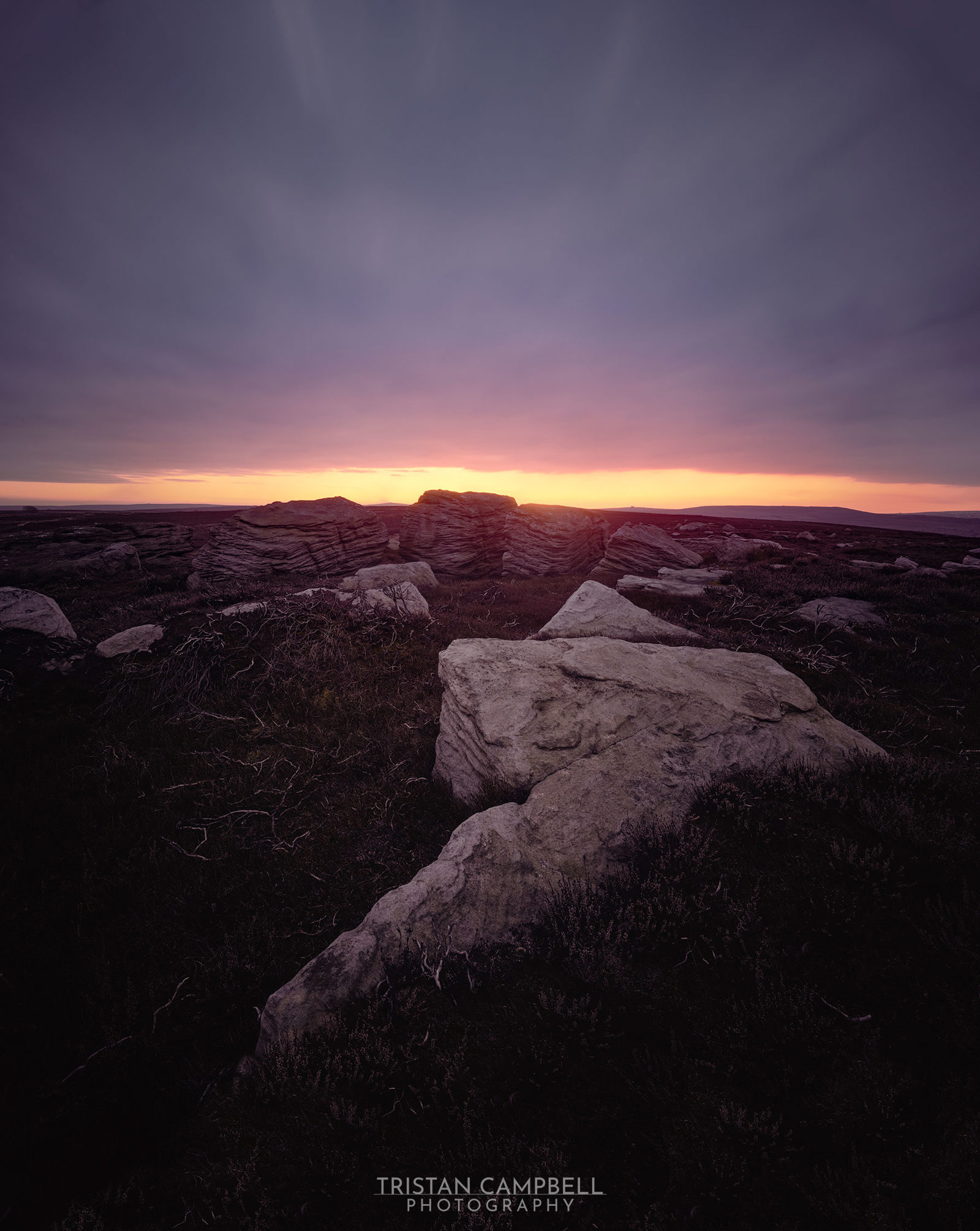 Rocky landscape at dusk with large stones in the foreground and a colourful horizon. The sky transitions from deep purple at the top to a bright orange near the horizon, suggesting a setting sun. The ground is covered in dark vegetation and scattered rocks, creating a moody, serene atmosphere.