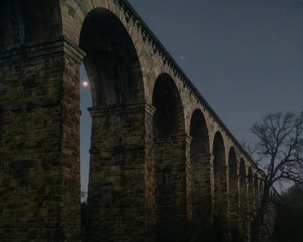 Stone railway viaduct arches at night with a blood moon glowing beneath one arch and stars above