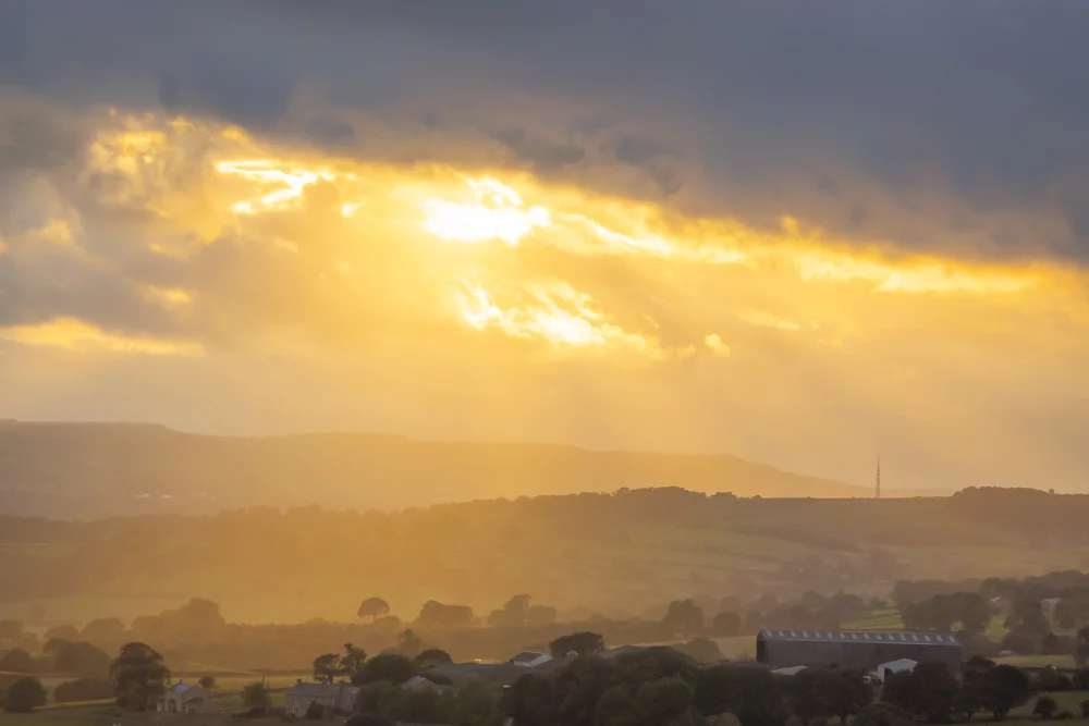 Sunbeams breaking through stormy cloud over a hazy moorland valley with farm buildings at dusk